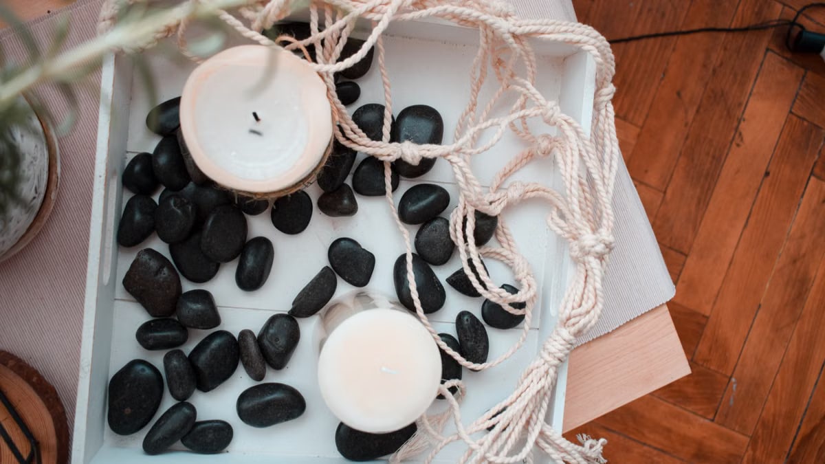 Top view composition with black decorative pebbles scattered around white candles on wooden tray with rope used for home decor in room with wooden floor